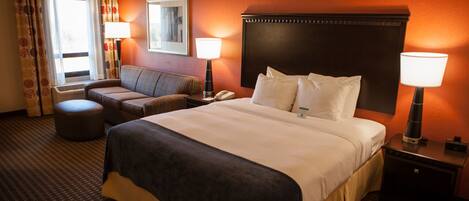 A hotel room with a large bed, a sofa, a nightstand, a lamp, and a window with curtains.