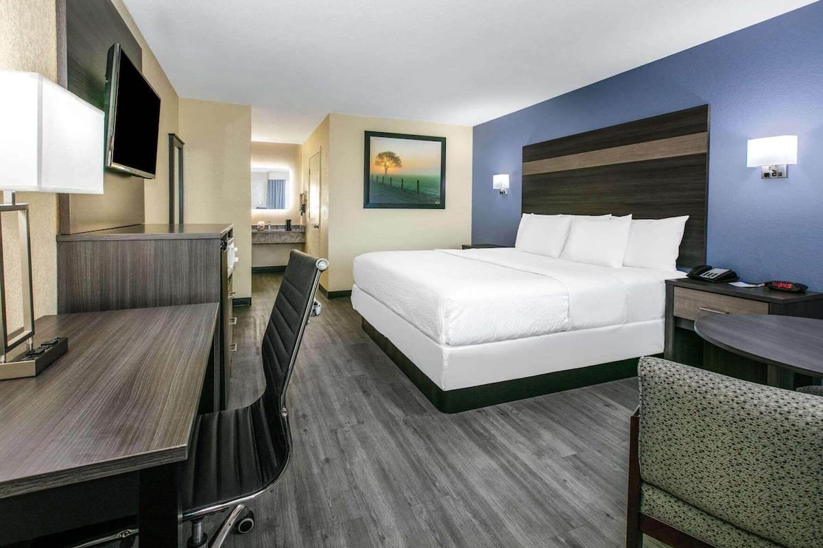 A hotel room with a bed, desk, chair, and a painting on the wall.