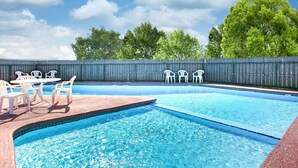 Une piscine entourée d’une clôture en bois et d’arbres, avec des chaises blanches et une table à proximité.