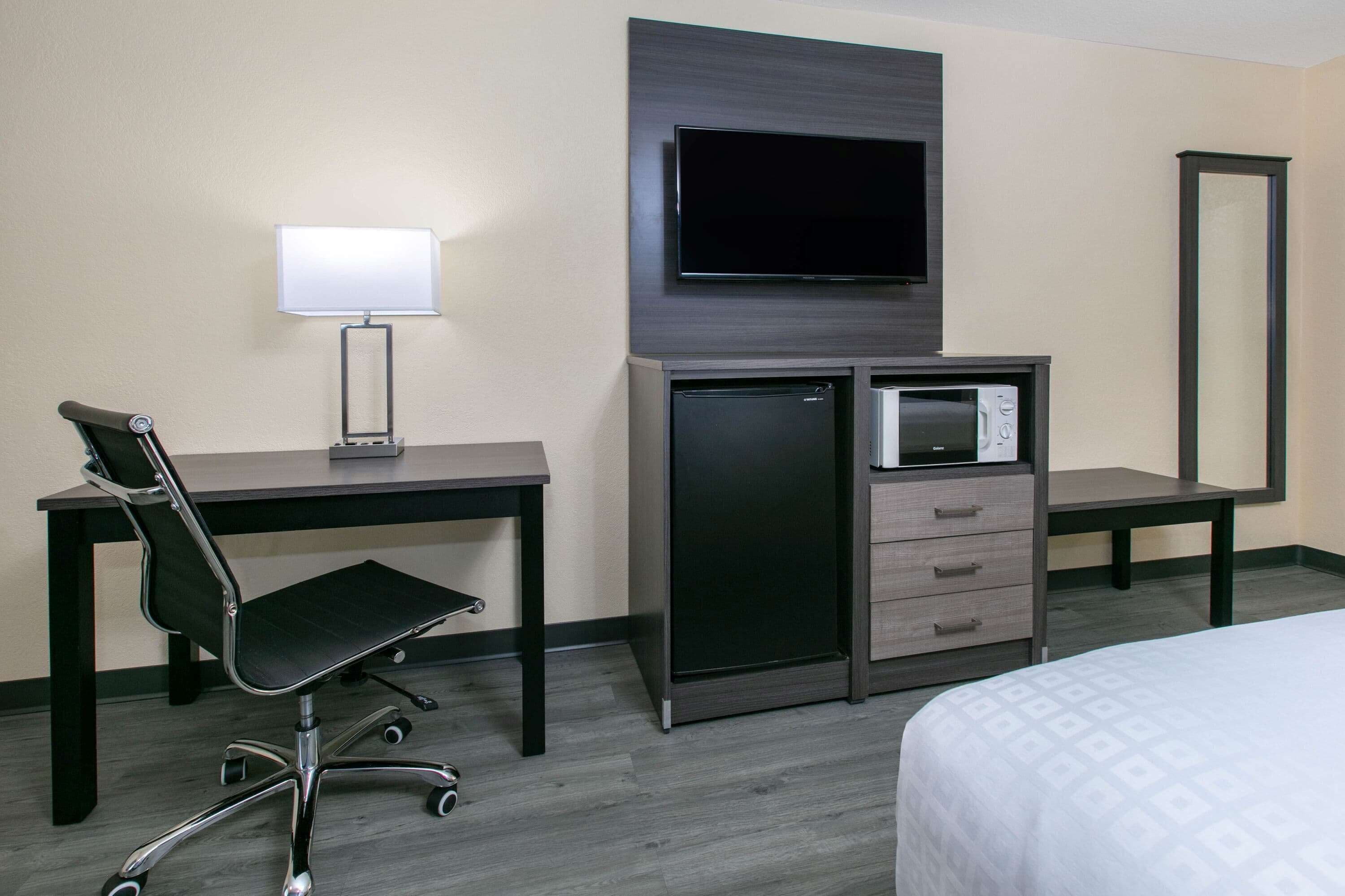 A hotel room with a desk, chair, TV, mini-fridge, and a bed.