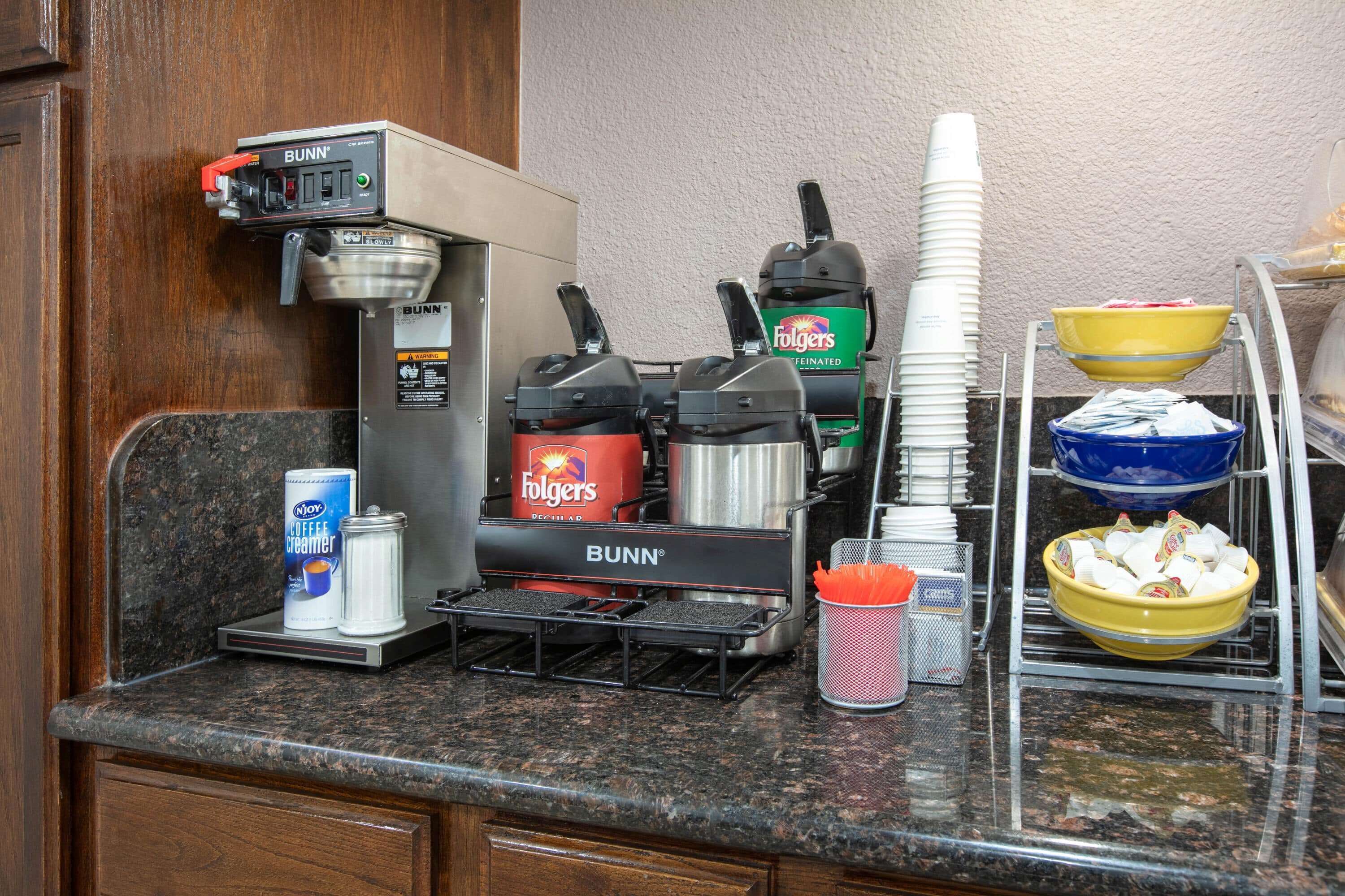 A kitchen area with a coffee machine, a coffee creamer dispenser, a Folgers coffee maker, and stacked paper cups.