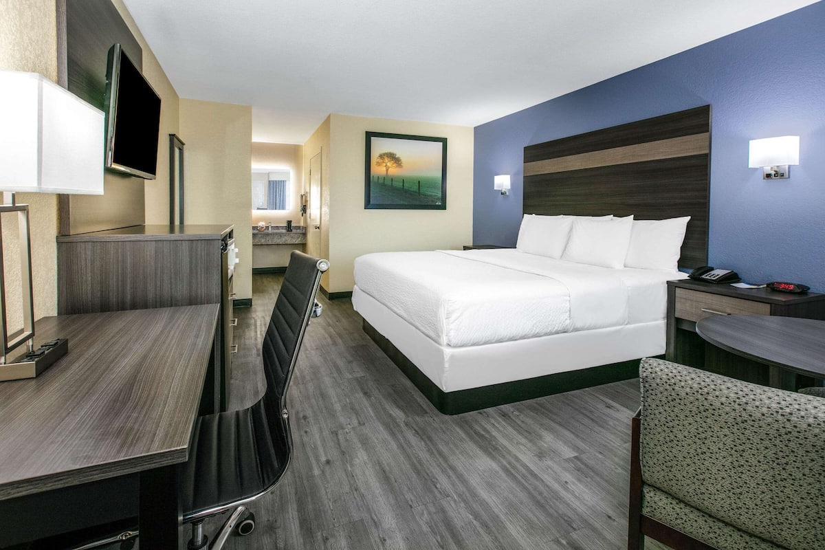 A hotel room with a bed, desk, chair, and a painting on the wall.