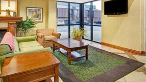 Een hotellobby met een groene bank, een gestreept fauteuil, een houten salontafel, en een groot raam met uitzicht op geparkeerde auto's en een gebouw.