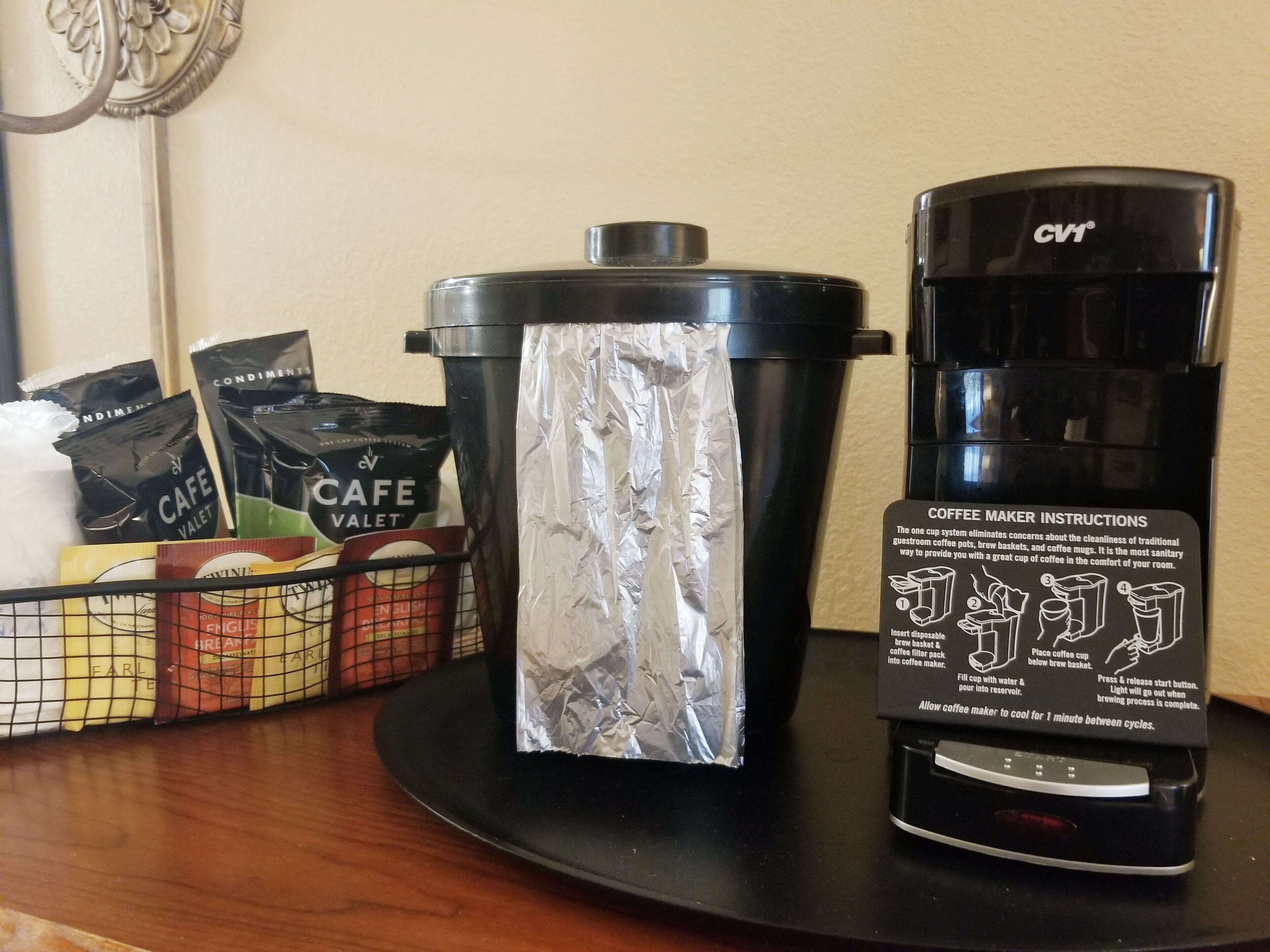 A coffee maker with instructions, a trash can wrapped in foil, and a basket of coffee pods on a table.