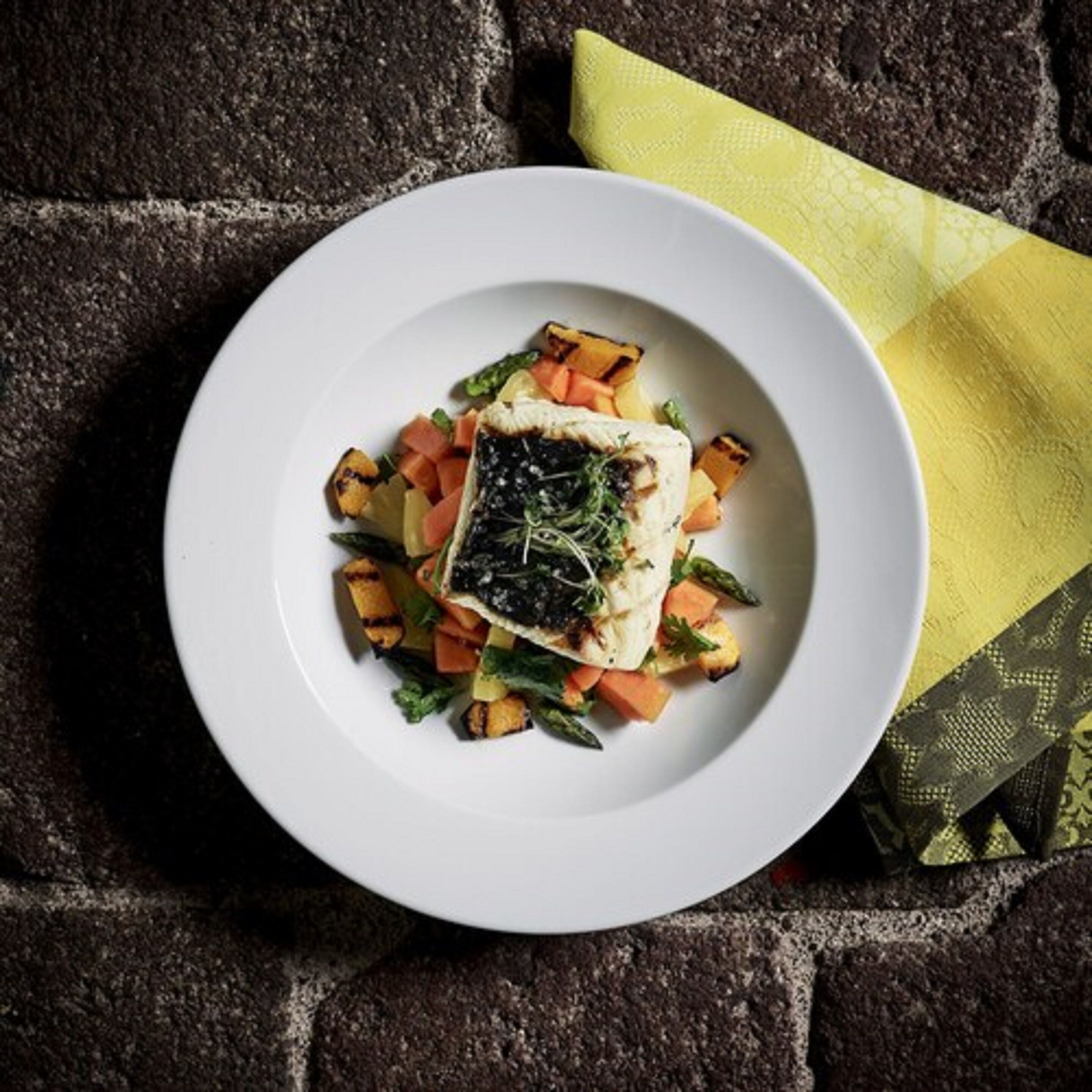 A white plate with a piece of fish, vegetables, and a yellow napkin on a textured surface.