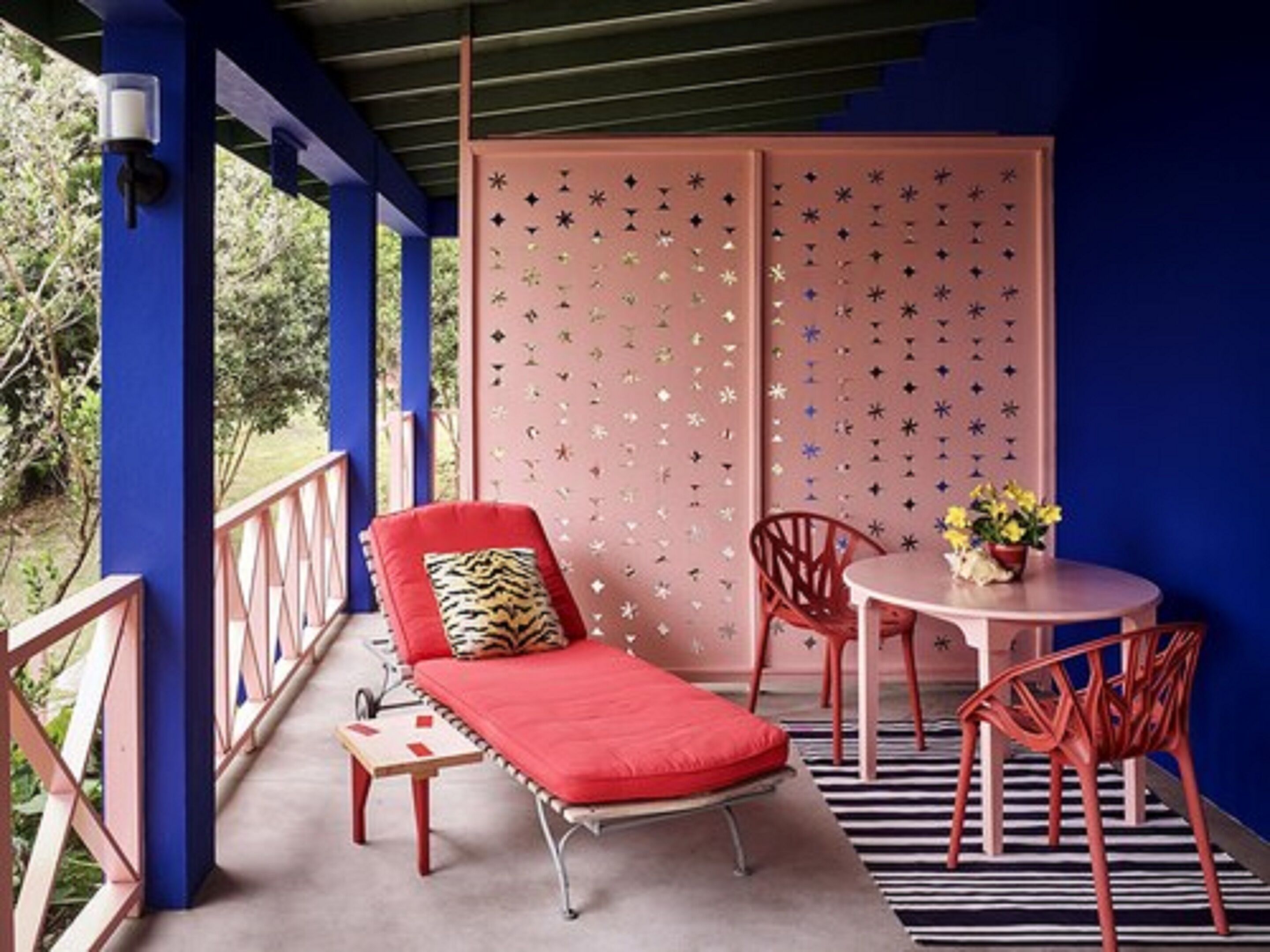 Un balcón con una silla lounge roja, una mesita con un jarrón de flores y una alfombra a rayas.