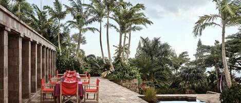 An outdoor dining area with a checkered floor, red tablecloths, and chairs, surrounded by palm trees and a small pool.