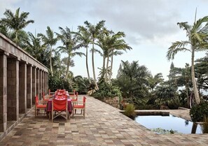An outdoor dining area with a checkered floor, red tablecloths, and chairs, surrounded by palm trees and a small pool.