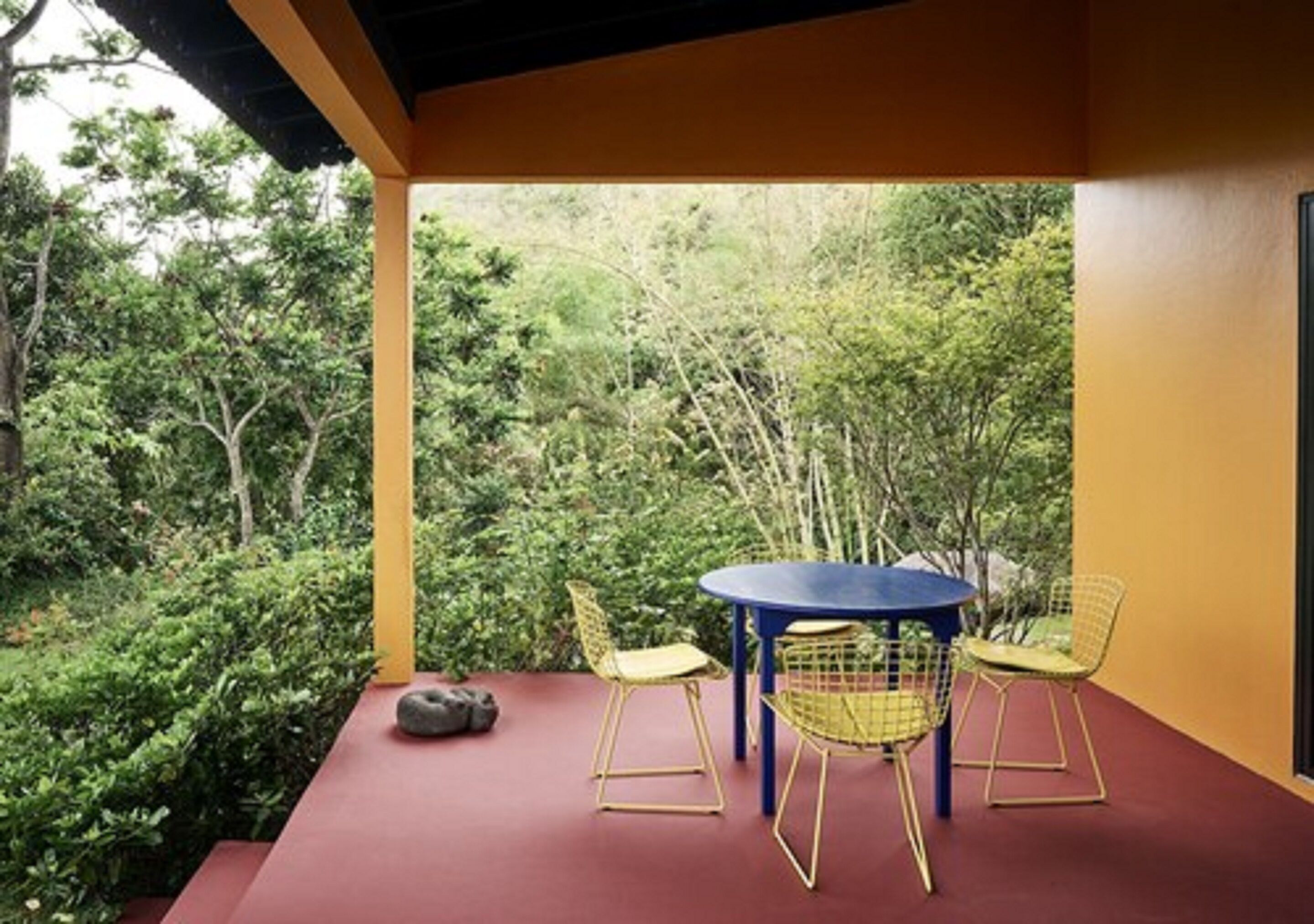 A covered outdoor seating area with a round table and two chairs, surrounded by lush greenery.