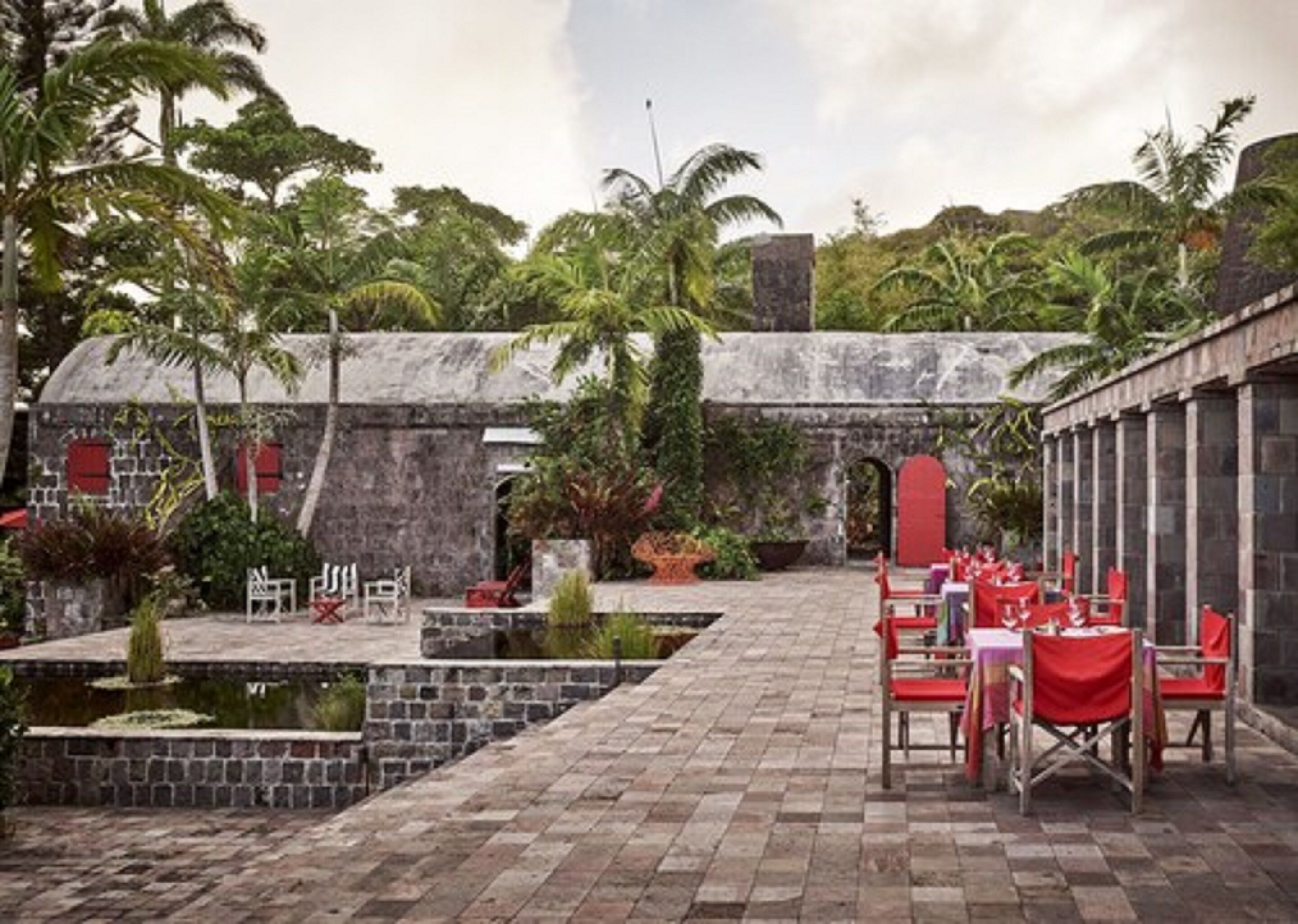 Un área de comedor al aire libre con paredes de piedra, palmeras y una fuente pequeña.