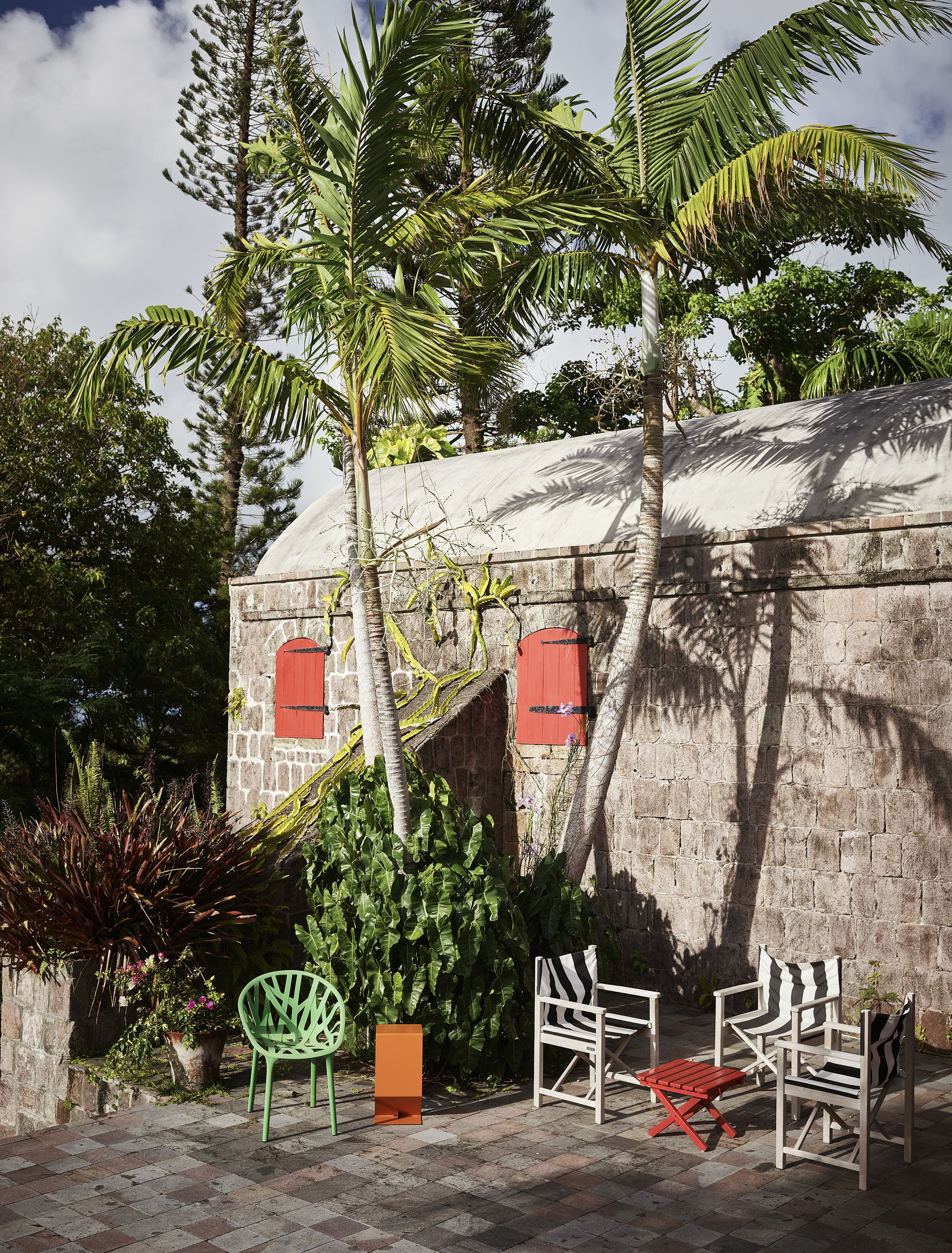 A stone building with red shutters, surrounded by lush greenery and palm trees.