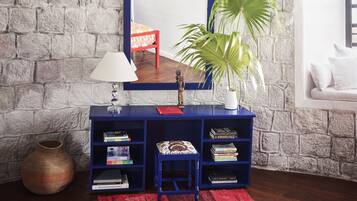 Quarto com parede de pedra, mesa consola azul com livros, cadeira vermelha, planta em vaso e sofá branco.