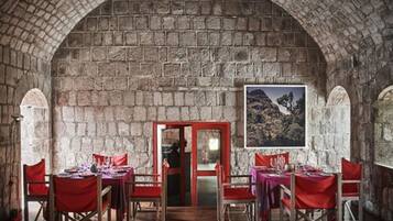 Un comedor de paredes de piedra con mesas y sillas rojas, piso de madera y un cuadro en la pared.