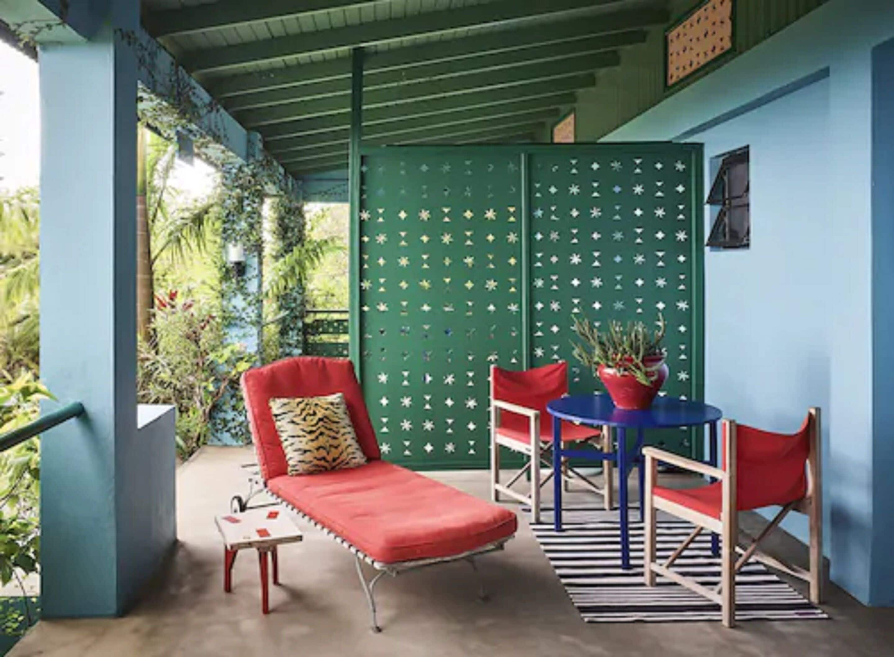 A covered outdoor seating area with a red lounge chair, two red chairs, a blue table, and a potted plant.