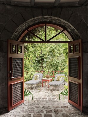 An open doorway leading to a patio with two striped chairs and a small table, surrounded by lush greenery.