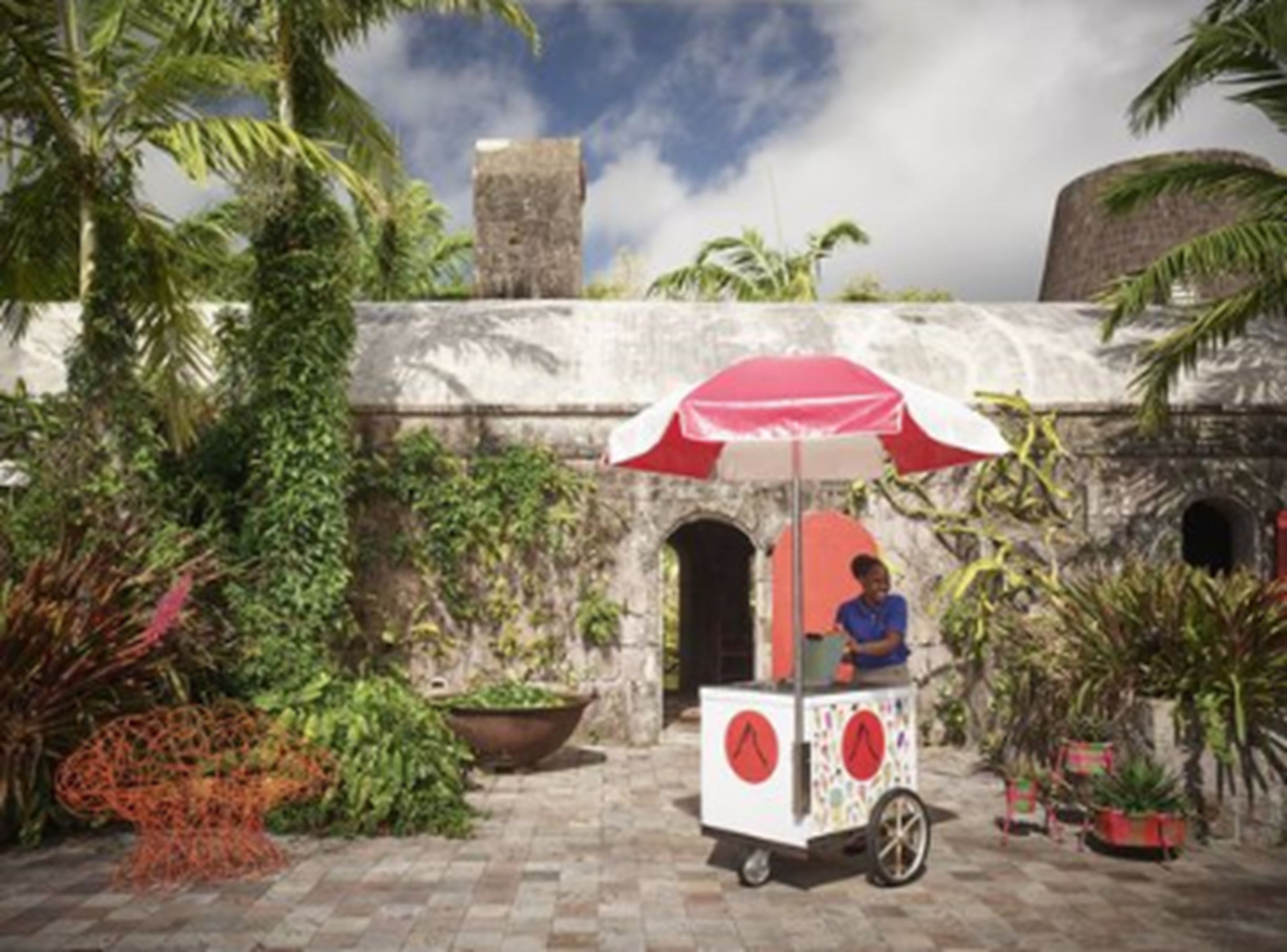 A vendor cart with a red umbrella in a courtyard with stone walls and tropical plants.