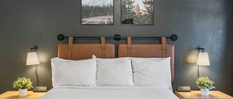 A hotel room with a bed, two bedside lamps, a nightstand with a plant, and two framed pictures on the wall.