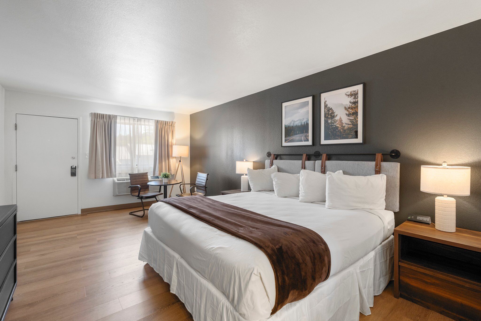 A hotel room with a large bed, a desk, a chair, a lamp, and two framed pictures on the wall.
