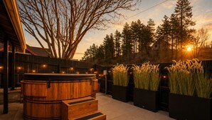 A wooden hot tub with steps, surrounded by tall grasses and trees, with a sunset in the background.