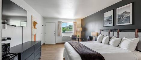 A modern hotel room with a large bed, a flat-screen TV, a desk with a lamp, and a window with curtains.