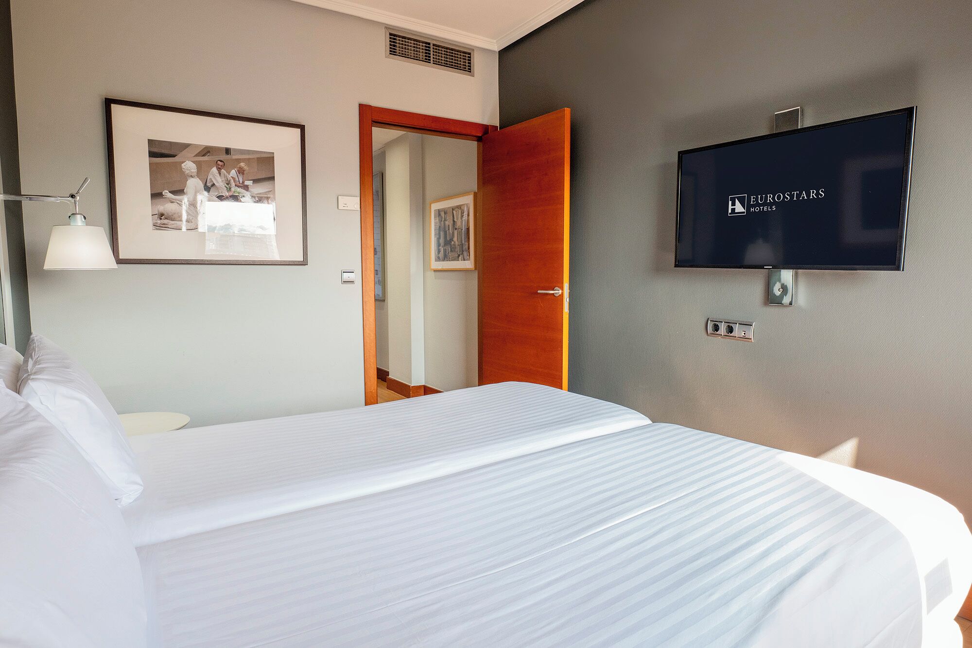 A hotel room with a bed, a TV mounted on the wall, a framed picture, and a door leading to another room.