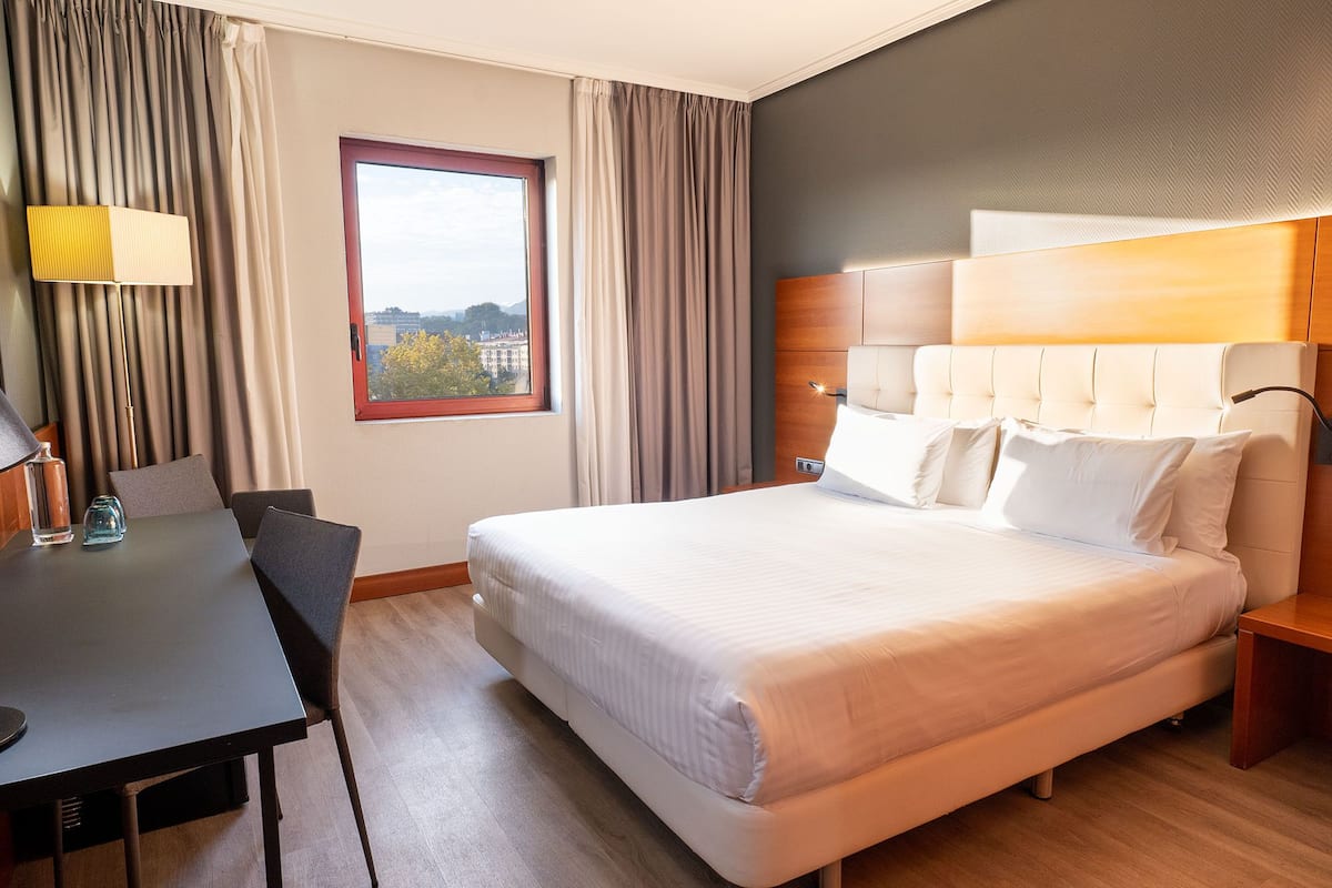 A hotel room with a large bed, a desk, a chair, a window with a view, and a lamp.