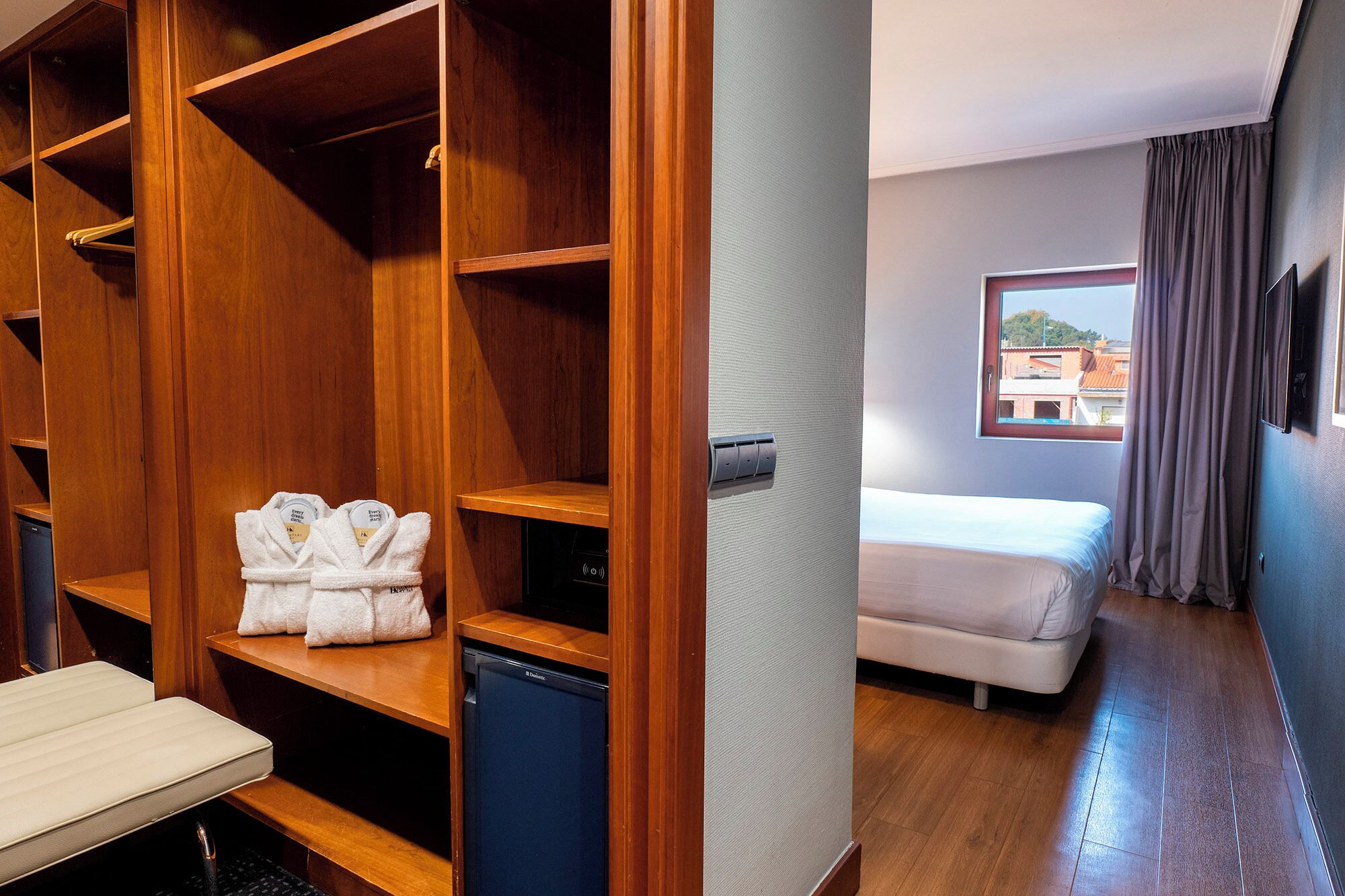 A hotel room with a wooden wardrobe, a small desk with a chair, a bed, and a window with a view of buildings.