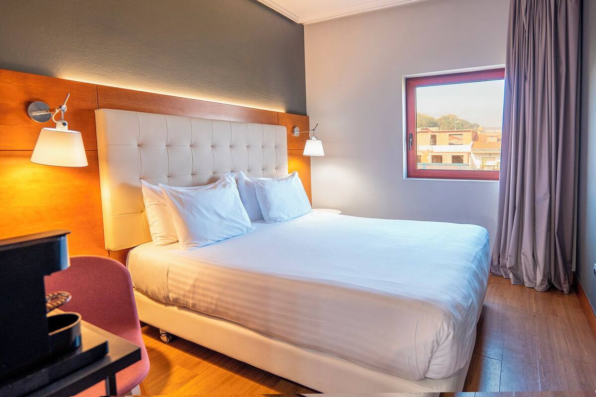A hotel room with a large bed, a wooden headboard, a desk with a chair, and a window with curtains.