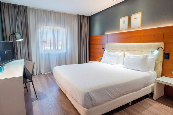 A hotel room with a large bed, a desk with a computer, a chair, and a window with curtains.