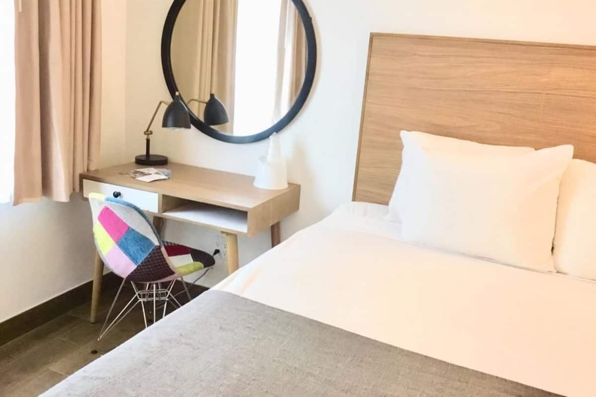Une chambre d’hôtel avec un lit, un bureau et une chaise, un miroir rond et une fenêtre avec des rideaux.