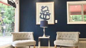 A room with two tufted armchairs, a side table with a lamp, a framed abstract artwork, and a window with a view of greenery. - San Diego Downtown Lodge (San Diego)