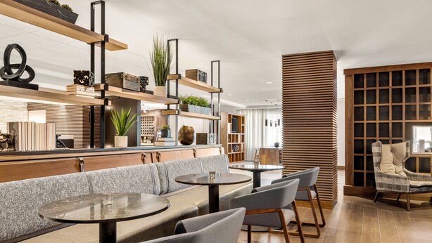 A modern cafe with round tables, grey cushioned seating, wooden shelves with plants, and a wooden accent wall.