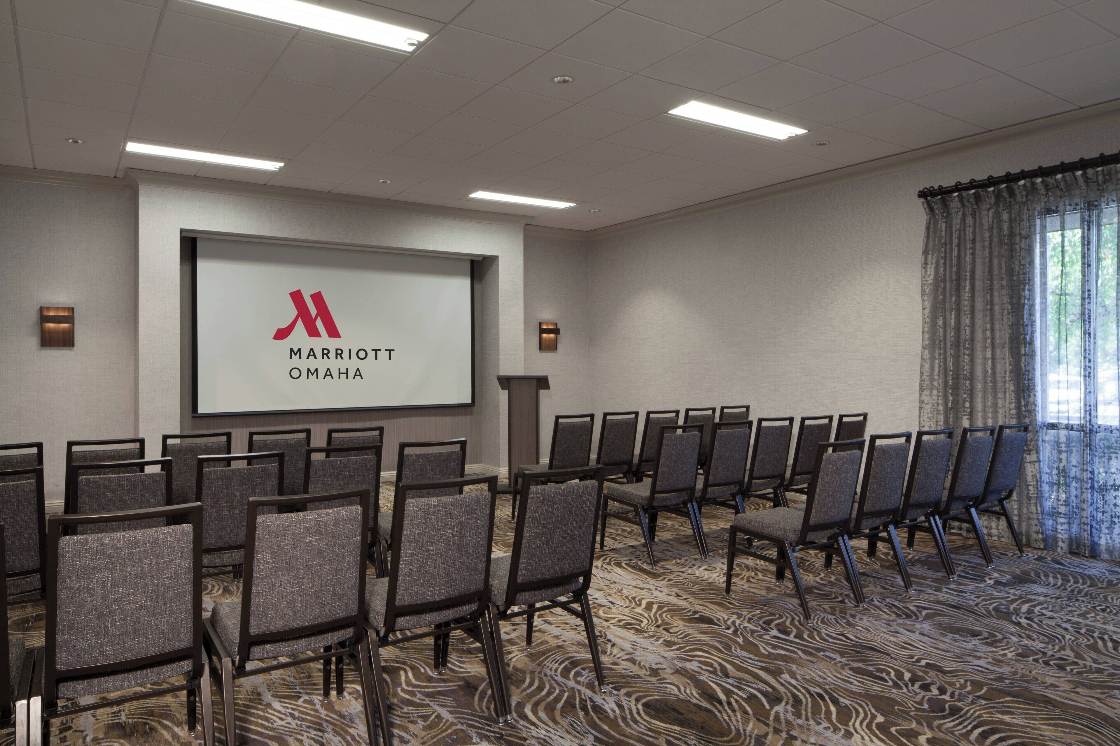 A conference room with rows of chairs, a large screen with 'Marriott Omaha' logo, and a carpeted floor.