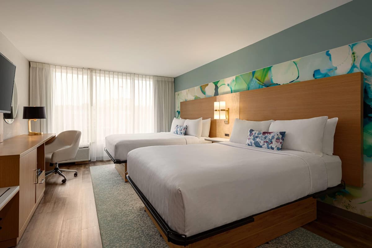 A hotel room with two beds, a desk, a chair, a TV, and a large abstract painting on the wall.
