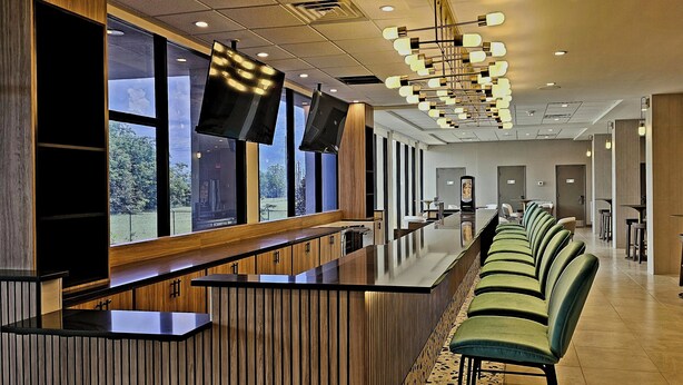 A modern hotel lobby with a long reception desk, green upholstered bar stools, and large windows offering a view of the outdoors. - Wyndham Garden Atlanta Airport (College Park)