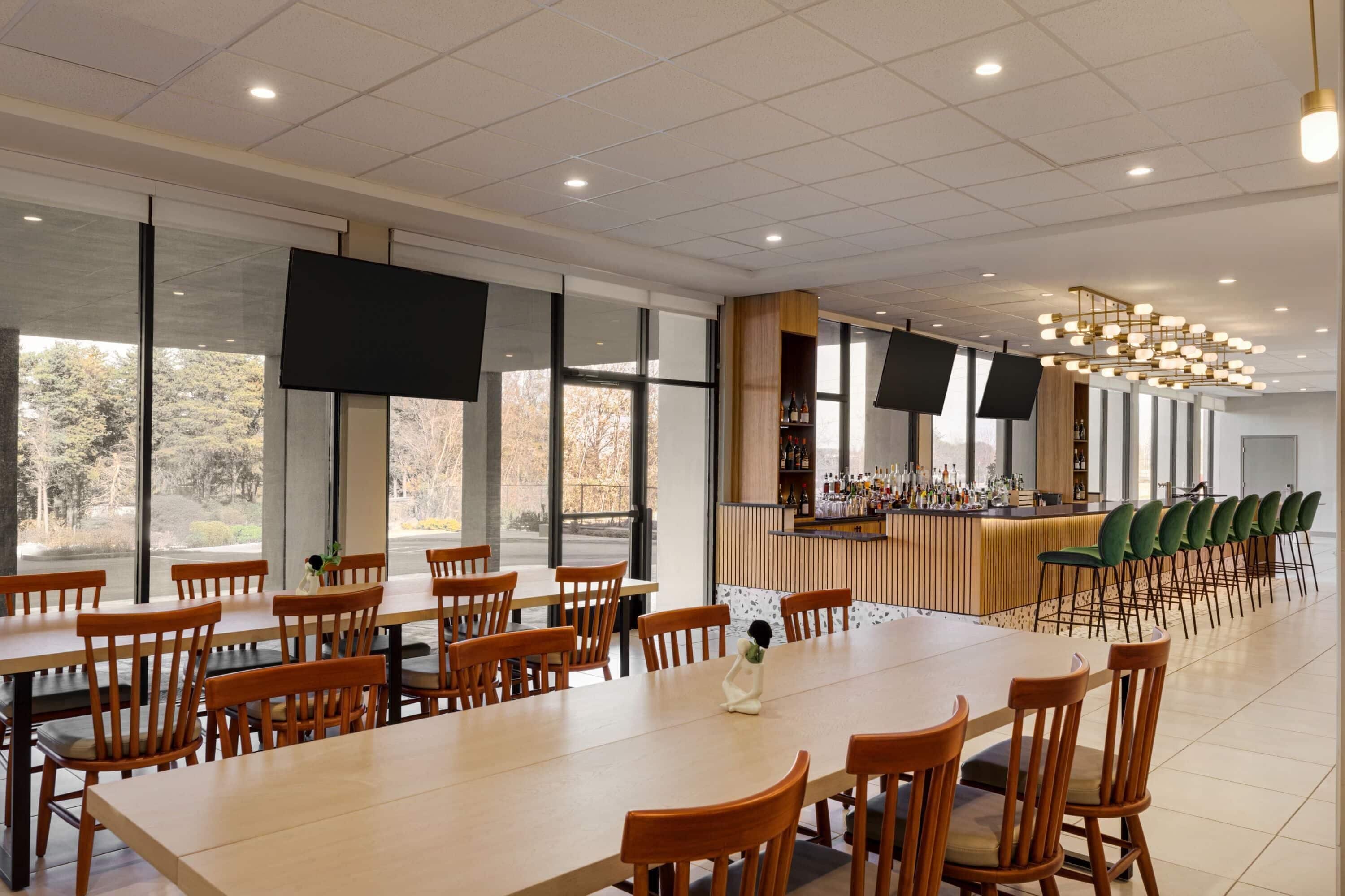 A spacious dining area with long wooden tables and chairs, a bar with a wooden counter, and large windows offering a view of trees.