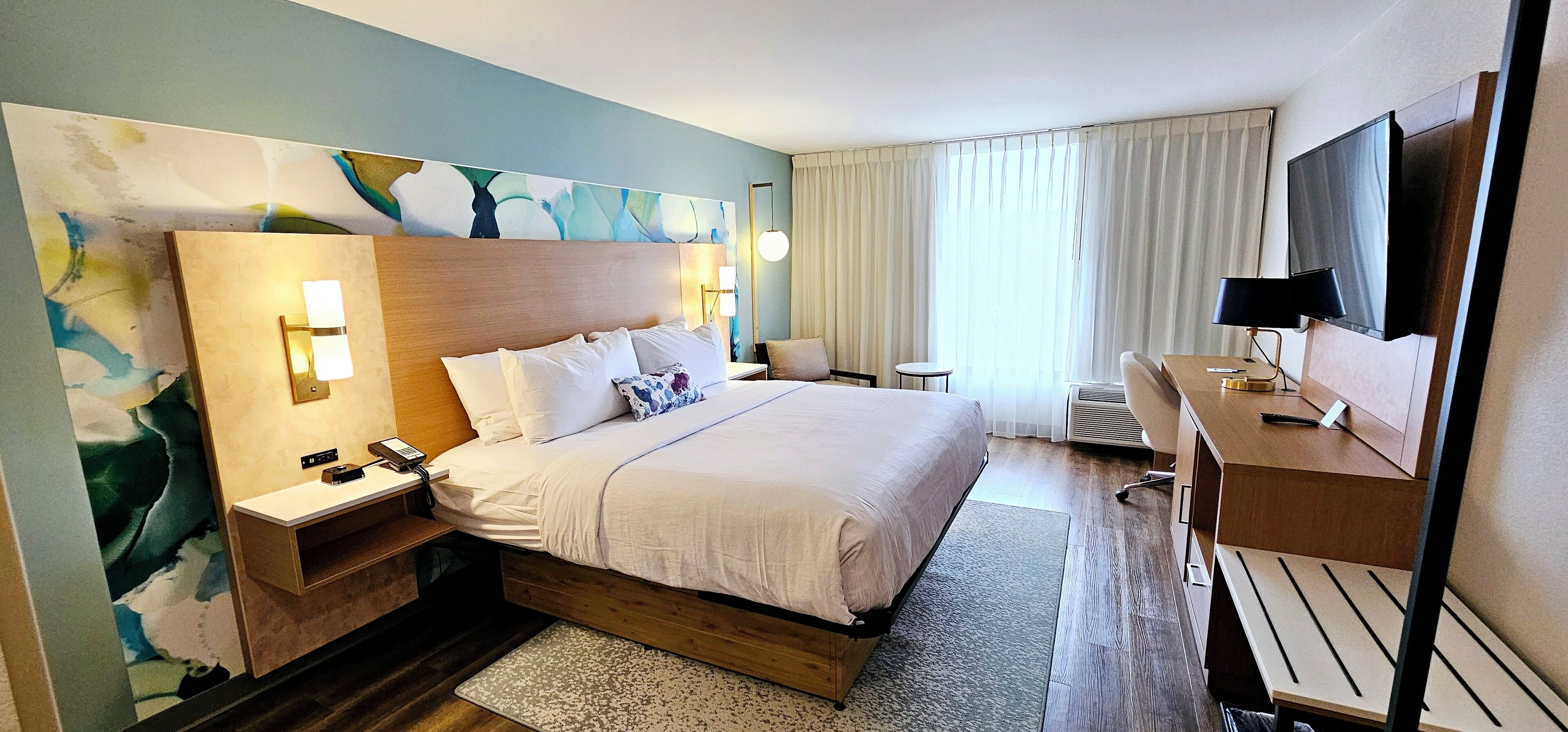 Une chambre d’hôtel avec un grand lit, des tables de chevet, un bureau avec une chaise, une télévision et une grande fenêtre avec des rideaux.