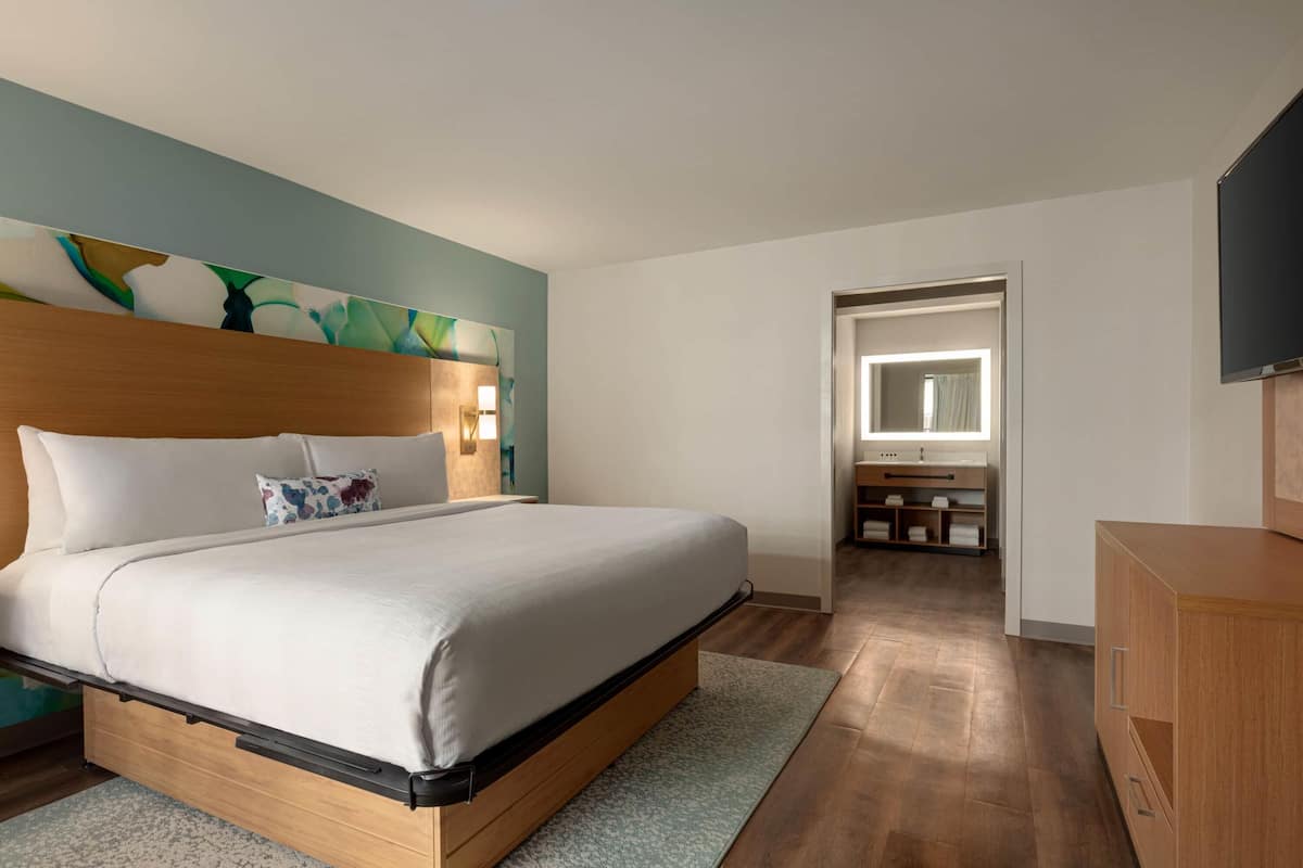 A modern hotel room with a large bed, a wooden headboard, a flat-screen TV, a bathroom with a mirror and sink, and a wooden cabinet.