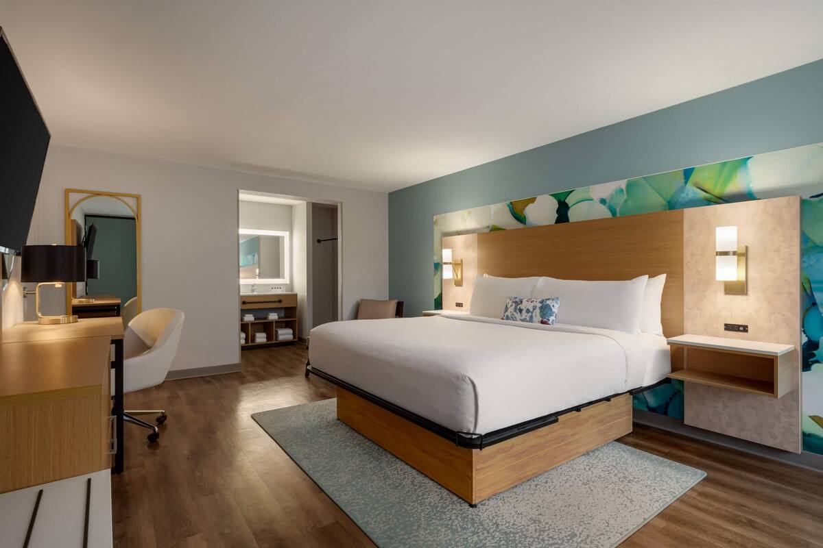 A modern hotel room with a large bed, a desk, and a chair. There is a bathroom visible through an open door.