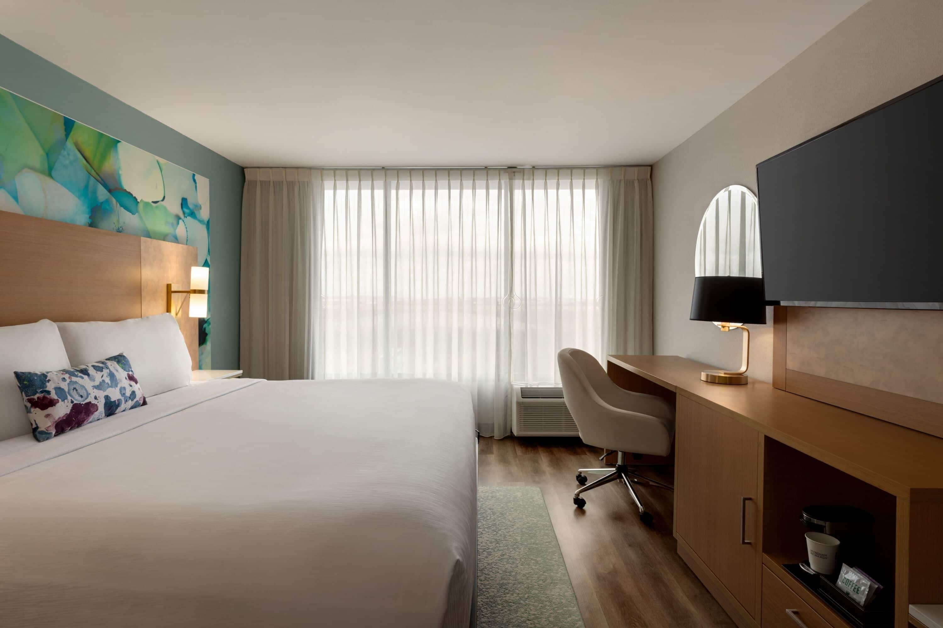 A hotel room with a large bed, a desk with a chair, a television, and a window with sheer curtains.