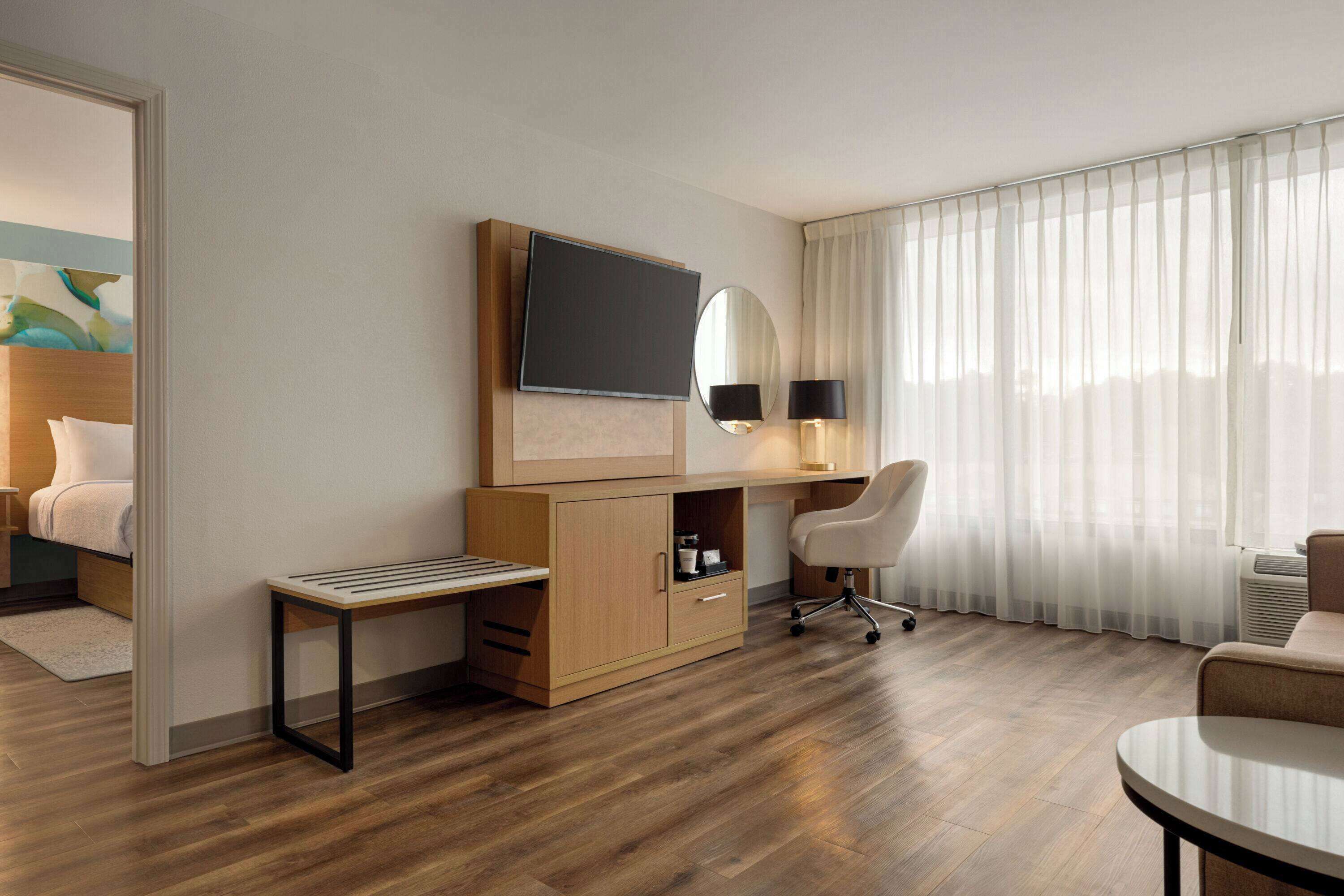 A hotel room with a wooden desk, a television mounted on the wall, a chair, a bed, and a large window with sheer curtains.
