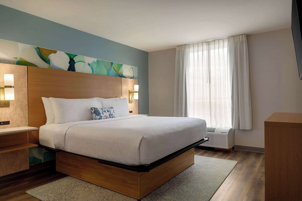 A modern hotel room with a wooden bed, a nightstand, a wall-mounted lamp, and a large window with sheer curtains.