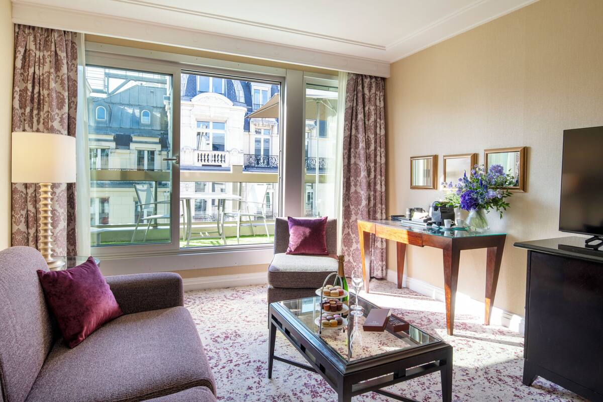 A modern hotel room with a large window, a sofa, a small table, and a TV.