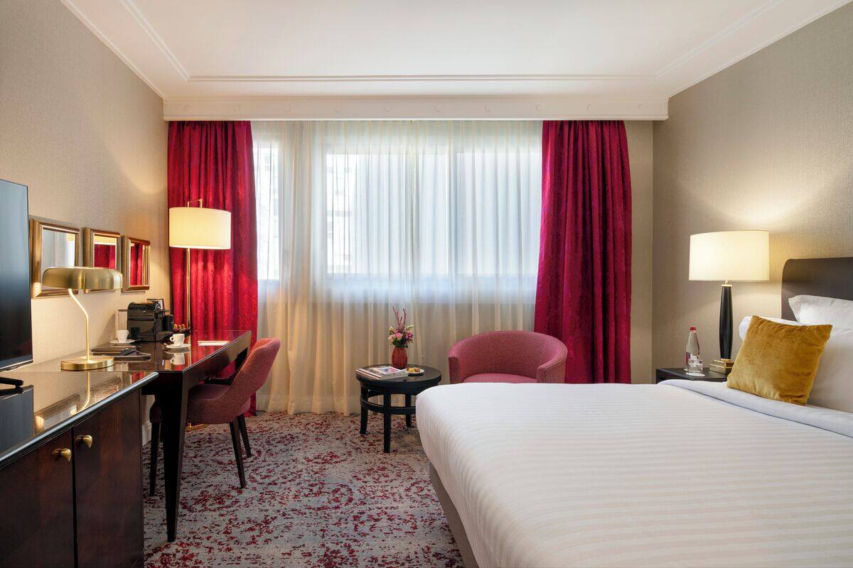 A hotel room with a bed, a desk, a chair, a TV, and a small table with a lamp.