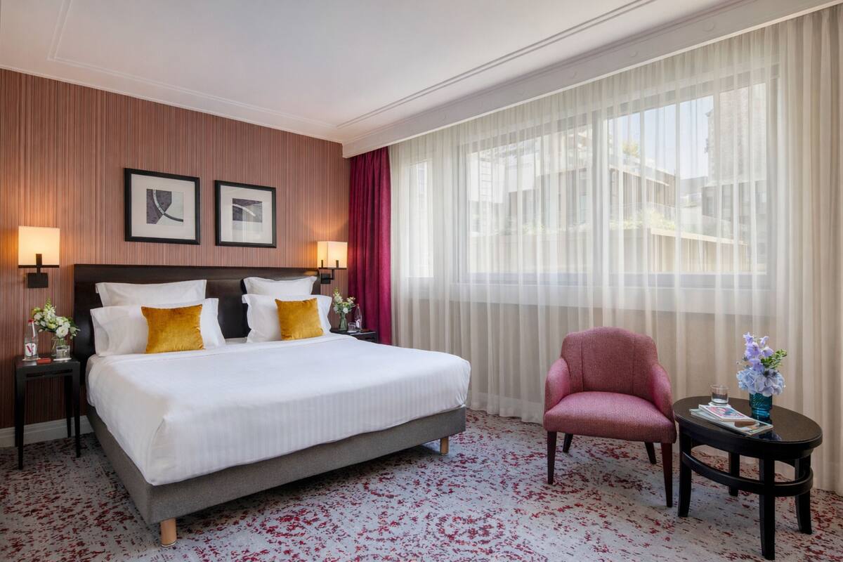 A hotel room with a large bed, two bedside tables with lamps, a chair, a small table with a vase, and a large window with sheer curtains.