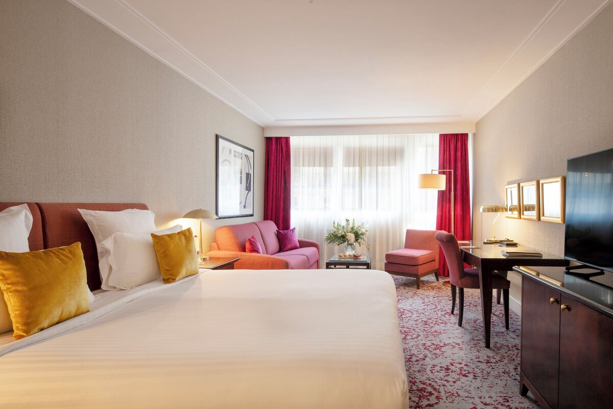 A hotel room with a large bed, a desk, a chair, a TV, and a window with curtains.