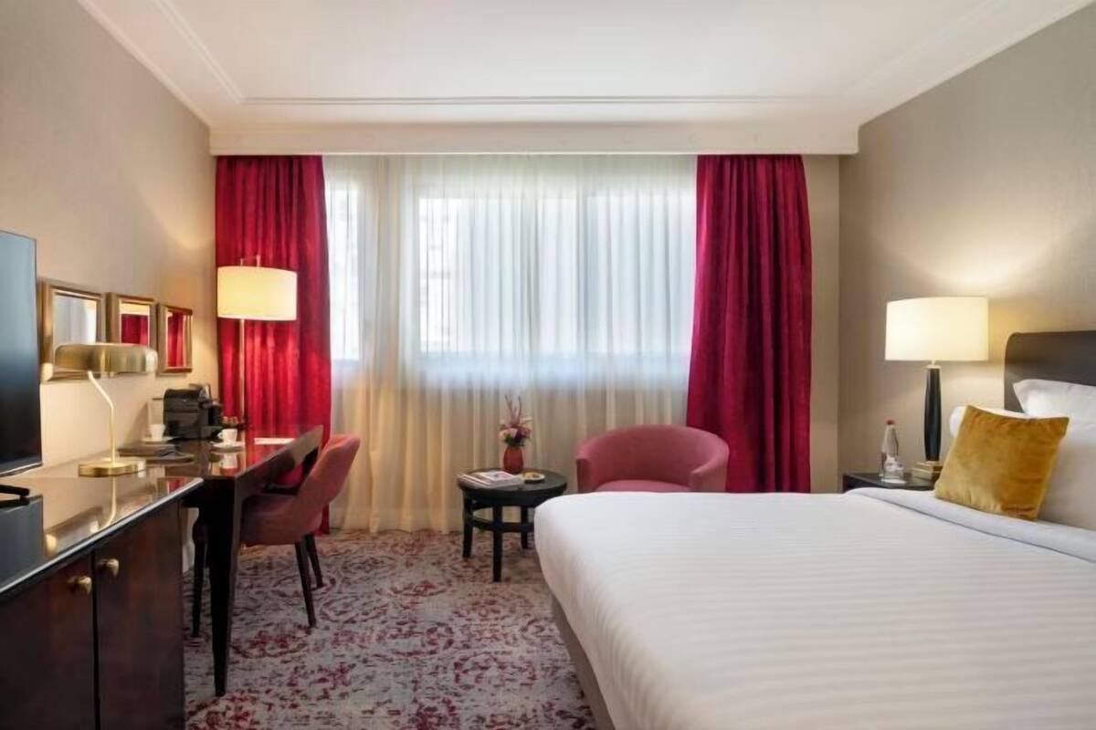 A hotel room with a bed, desk, chair, and a small table with a lamp.