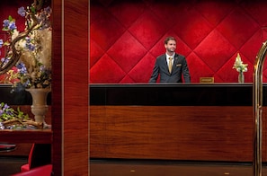 A reception desk with a man standing behind it, a red velvet wall, and a vase with flowers. - Warwick Champs-Elysées Paris (Paris)