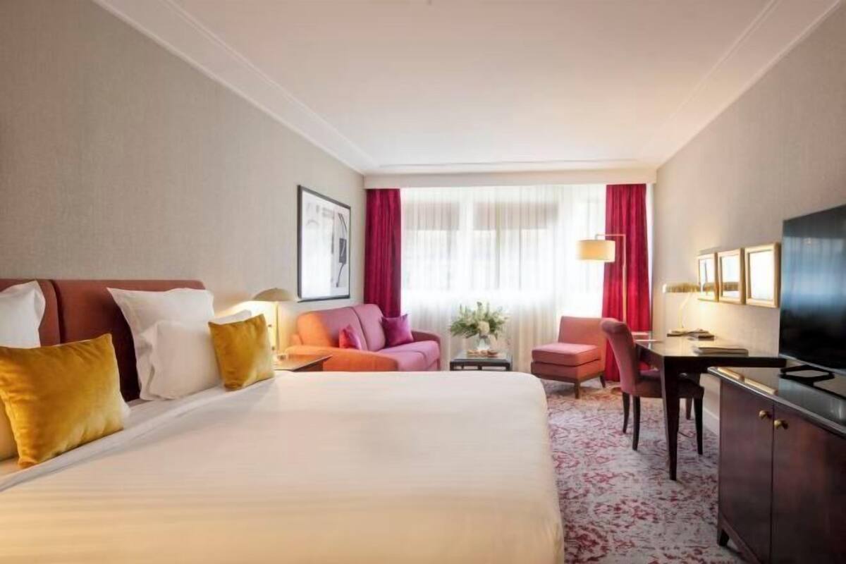 A hotel room with a large bed, a desk, a chair, a TV, and a window with curtains.