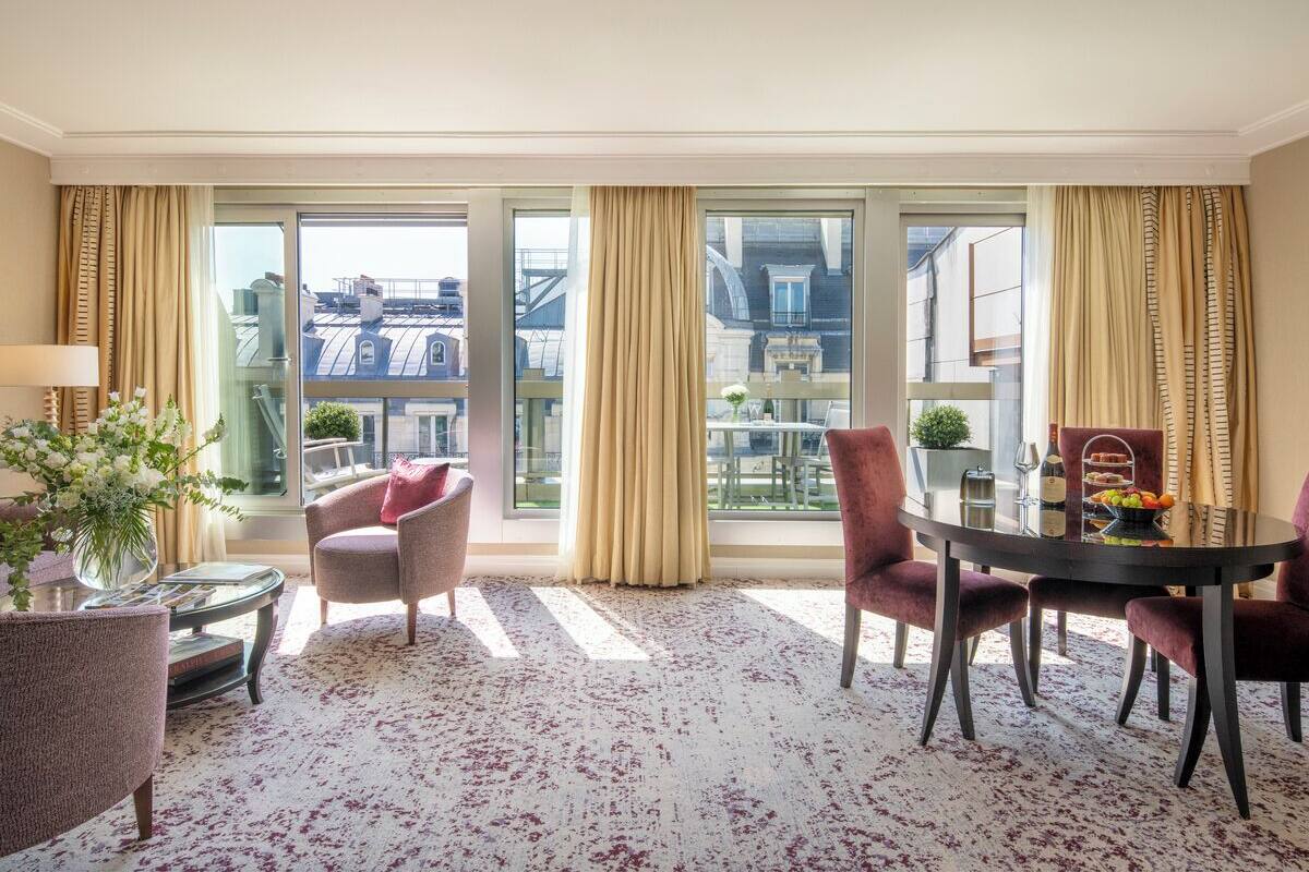 A hotel room with a large window, a round dining table, two armchairs, a small side table, and a vase with flowers.
